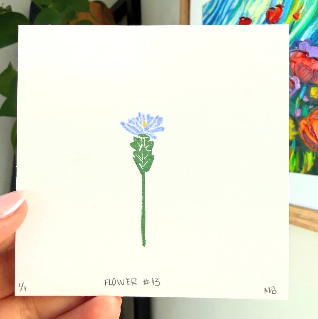 Hand holding a small card with a drawing of a flower against a colorful painting background.